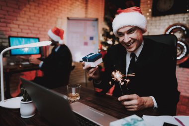 Yeni yıl arifesinde ofiste kırmızı şapkalı bir iş adamı. Ofiste Noel ağacı. Laptop üstünde okul sırası. İş kavramı. Siyah elbiseli adam. Yeni yıl kutlama. Dijital aygıt kullanarak. Hediye kutusu.