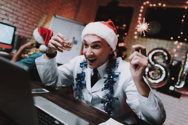 年轻的医生在红帽办公室在新年前夕 在办公室的圣诞树 电脑在书桌上 圣诞老人帽里的人穿白大衣的人 庆祝新年 使用数字设备 — 图库照片
