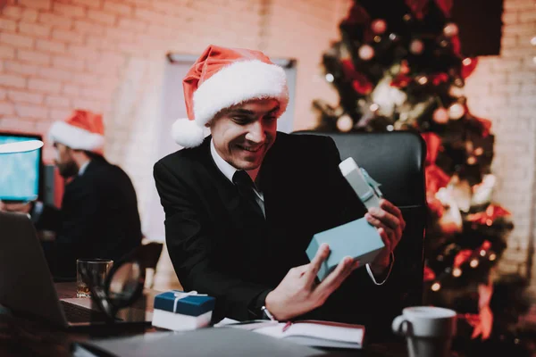 Businessman in Red Cap in Office on New Year Eve. Christmas Tree in Office. Laptop on Desk. Business Concept. Man in Black Suit. Celebrating of New Year. Using Digital Device. Gift Box.