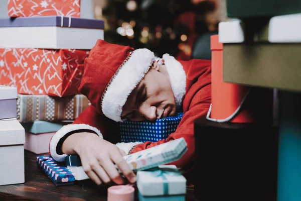 圣诞老人服装与礼品盒的年轻人 新年前夜 在办公室的圣诞树 庆祝新年 红色的服装 红帽人成堆的箱子 人睡在桌上 — 图库照片