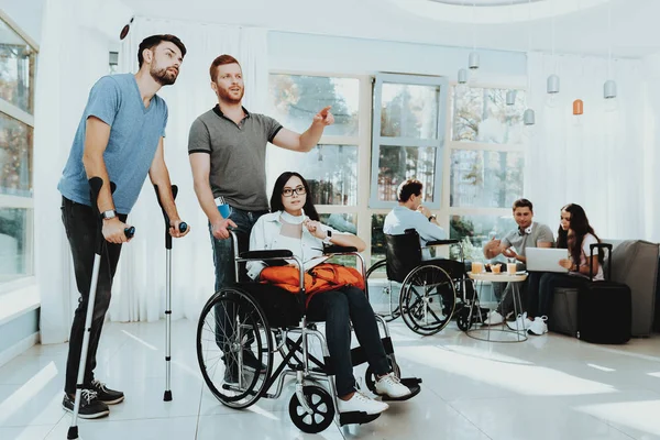 People in Wheelchair. Disabled in Hall. Woman in Wheelchair. Man on Crutches. Room with Panoramic View. Gray Sofa. White Interior. Meeting Disabled. Limited Opportunities. Man with Woman in Wheelchair