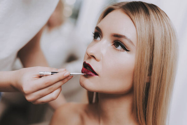 Blond Girl in Beauty Salon. Makeup in Beauty Salon. Woman Colors Lips. Woman and Makeup Artist. Beautiful Woman. Blond Girl with Shining Skin. Pretty Blond Girl. Model and Makeup Artist. Red Lips.
