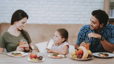 Cam süt olan kız. Aile kahvaltı yapıyor. Aile masada gülümseyerek. Meyve plaka üzerinde. Sandviç plaka üzerinde. Beyaz tabak. Cam suyu. Gıda üzerine plaka. Mutlu bir aile. Gülümseyen insanlar. Beyaz iç.