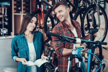 Kız bisiklet danışman gibi seçmeyin. Denim giysileri güzel kızı kareli gömlek ve gülüyor spor mağazasında giymiş yakışıklı satıcı ile ayakta döngüsü satın alma hakkında şüpheci görünüyor