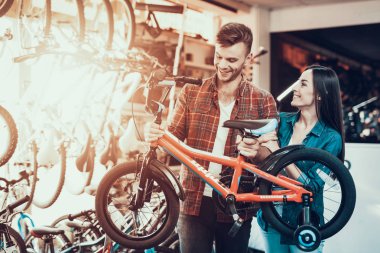 Genç çift çocuklar bisiklet deposunda seçer. Yakışıklı adam kareli gömlek tutan çocuk bisiklet el ve gülüyor giymiş güzel kız spor döngüsü dükkanda ayakta bakar