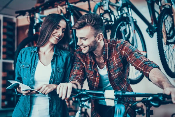 幸せな若いカップルが話している間は新しい自転車を選択します デニムで美しい少女服購入サイクル スポーツ店で市松模様のシャツと笑を着てハンサムなコンサルタントと話しています — ストック写真