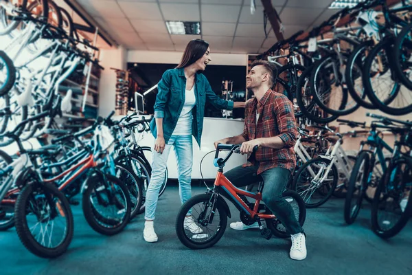 幸せな若いカップルは 子供自転車を選択します デニムの服スポーツ サイクル ショップに立っている美しい少女を見て子供の自転車と笑いに座っている格子縞シャツを身に着けているハンサムな男 — ストック写真