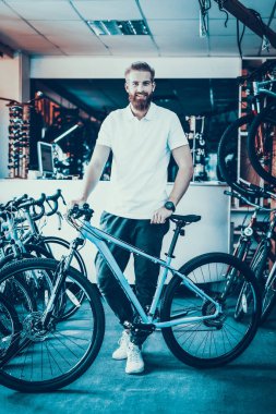 Satıcı pozlar bisiklet spor salonu içinde yakınındaki gülümseyerek. Neşeli sakallı beyaz erkek satıcı kameraya bakarak beyaz T-Shirt giyen portresi. Mutlu danışman tutar döngüsü spor mağazasında