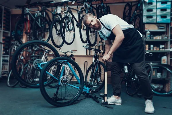 Guy Pumps Bicycle Wheel with Air in Bike Workshop. Portrait of Young Bearded Bicycle Mechanic Wearing White T-Shirt and Apron Examines and Fixes Modern Cycle. Bike Maintenance Concept