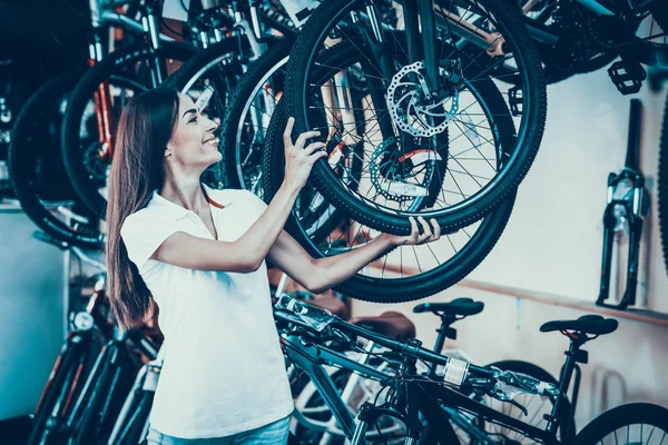 Beautiful Young Woman Shows Row of Modern Bicycles. Portrait of Cheerful Stylish Caucasian Female Seller Wearing White T-Shirt Holds Cycle Wheel. Happy Consultant in Cycle Sport Store
