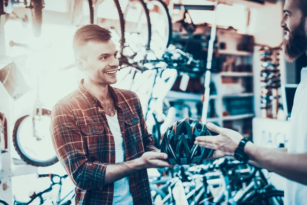 Man Look for Helmet for Bike Rides in Sport Store. Young Caucasian Smiling Guy Wearing Red Checkered Shirt Buys Modern Helmet Talks with Bearded Shop Assistant in Bicycle Shop