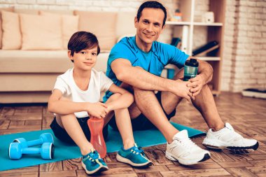 Çocuklar için spor. Baba ve çocuk tespit yapmak. Evde spor. Sağlık kavramı. Sağlıklı bir yaşam tarzı. Beyaz kanepe. Katta oturuyor. Erkek eğitmen. Elinde şişe su. Eğitimden sonra dinlenme. Minderin üstüne otur.