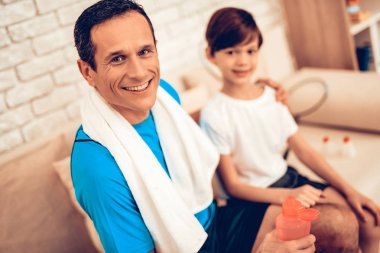 Çocuk elinde su ile. Baba ve çocuk tespit yapmak. Evde spor. Sağlık kavramı. Sağlıklı bir yaşam tarzı. Beyaz kanepe. Erkek eğitmen. Elinde şişe su. Eğitimden sonra dinlenme. Oturun.