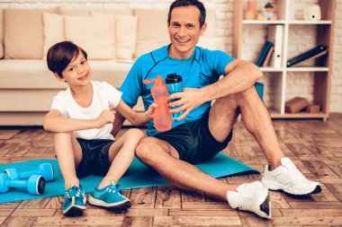 Çocuklar için spor. Baba ve çocuk eğitim sonrası. Evde spor. Sağlık kavramı. Sağlıklı bir yaşam tarzı. Beyaz kanepe. Katta oturuyor. Erkek eğitmen. Elinde şişe su. Eğitimden sonra dinlenme. Minderin üstüne otur.