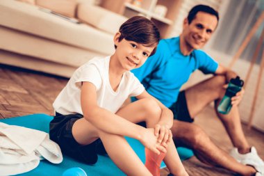 Çocuklar için spor. Baba ve çocuk tespit yapmak. Evde spor. Sağlık kavramı. Sağlıklı bir yaşam tarzı. Beyaz kanepe. Katta oturuyor. Erkek eğitmen. Elinde şişe su. Eğitimden sonra dinlenme. Minderin üstüne otur.