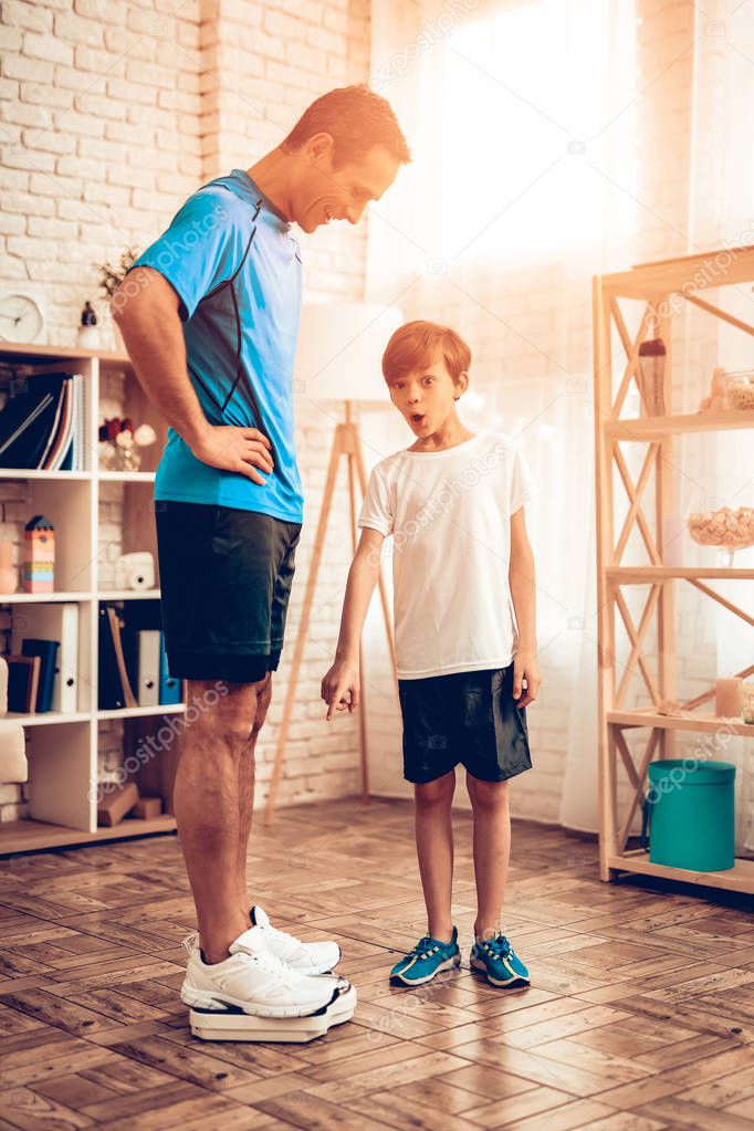 Man Standing on Scales. Father and Son do Spotting. Sport at Home. Warm Up in Quarter. Gymnastic Mat. Doing Sports. Man and Boy Train at Home. Man in Sports Uniform. Athletes in Apartment.