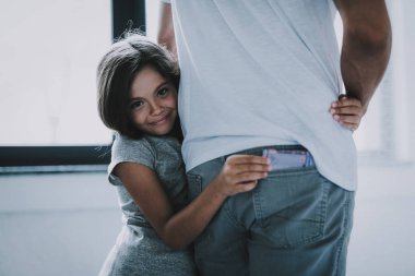 Girl Hugs Father Steals Money From Jeans Pocket. Smiling Little Caucasian Girl Standing Indoors Thieves Guietly Pulls Out Father's Cash from Back Pocket. Difficult Child Concept clipart