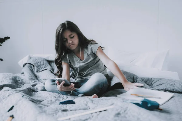 Little Girl Listens Music and Draws in Bedroom. Frowning Cute Black-Haired Child Holds Smartphone Sits on Large Gray Bed in Modern Apartment Uses White Earphones. Leisure Time Concept