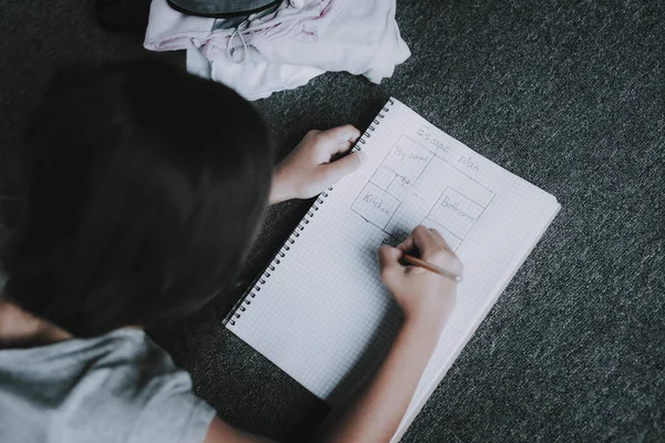 メモ帳で床の上に横たわる少女の平面図を描画します 灰色のカーペットの上に横たわる黒髪の子供のポートレート クローズ アップ 白いノートに鉛筆で書き込みます レジャー時間の概念 — ストック写真