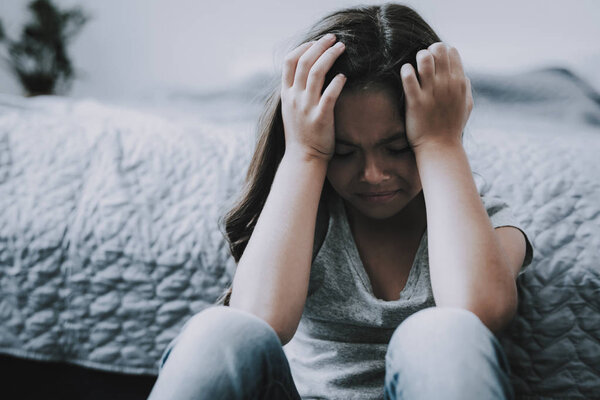 Плачущая маленькая девочка сидит на полу в помещении. Крупный план Портрет напуганного несчастного черноволосого белого ребенка с закрытыми глазами, сидящего у кровати в серой футболке, держит руки за головой
.