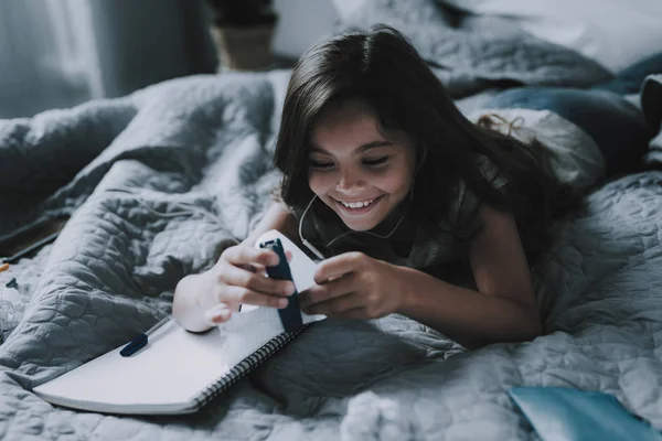 快乐的女孩听音乐在耳机在卧室 微笑的黑发孩子穿着休闲衣服使用蓝色订书机躺在大灰色床与记事本 休闲时间概念 — 图库照片