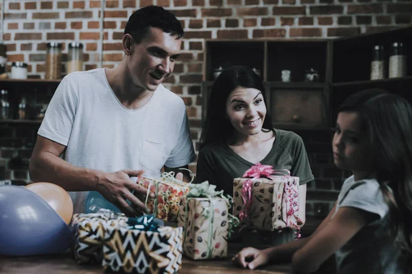 Parents Give Presents to Indifferent Daughter. Smiling Mother and Father Congratulate Girl with Bright Gifts on Birthday in Modern Kitchen in Wooden Style. Daughter Feels Bored and Looks away