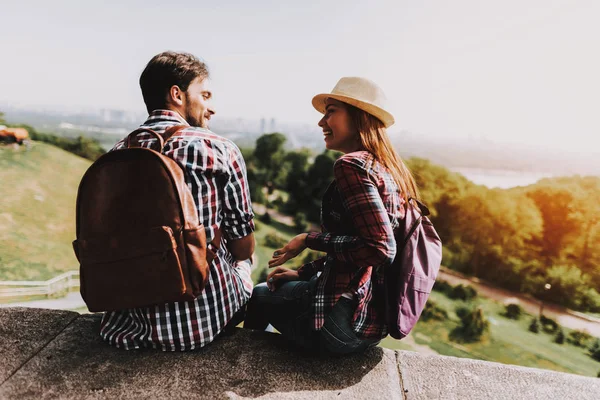 Young Couple Sitting on Ledge and Looking at Park. Young Man and Woman with Backpacks Sitting Together on Concrete Ledge. Couple Enjoying Beautiful View of Nature. Traveling Concept