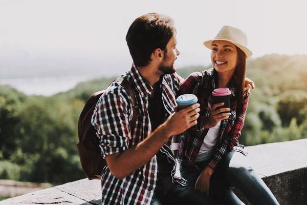 Happy Couple Sitting on Ledge and Drinking Coffee. Young Man and Woman with Backpacks Sitting Together on Concrete Ledge. Couple Enjoying Beautiful View of Nature. Traveling Concept