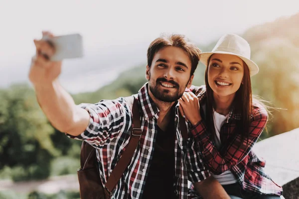 Couple Sitting on Concrete Ledge taking Selfie. Young Man and Attractive Woman with Backpacks Sitting Together on Concrete Ledge. Couple Enjoying Beautiful View of Nature. Traveling Concept