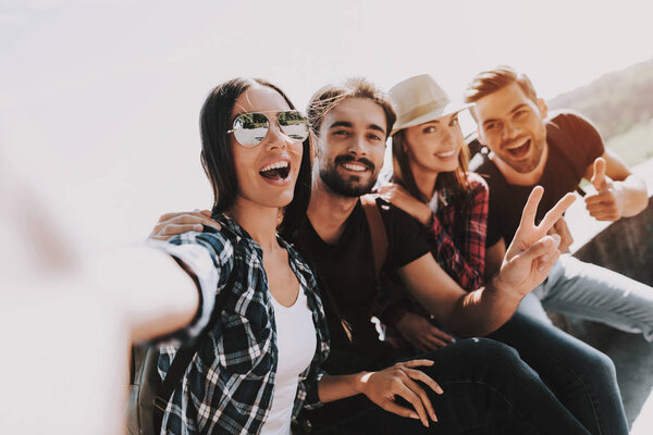 Young Smiling People Sitting in Park taking Selfie. Group of Young Friends with Backpacks Sitting Together taking Selfie by Using Smartphone. Travelers Enjoying Summer. People and Friendship Concept