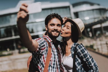 Selfie alarak sırt çantaları ile genç mutlu çift. Yakışıklı adam ve güzel kadın alarak Selfie yanında beyaz Smartphone kullanarak cam bina önünde. Yaz tatili zevk genç gezginler