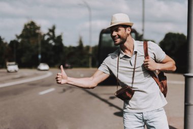 Genç Turizm yolda otostop sırt çantası ile. Kayıtsızca giyinmiş kamera ile yakışıklı adam karayolu üzerinde araç yakalamak için boyun. Turizm ve insanlar kavramı. Aktif yaşam tarzı. Yaz tatili
