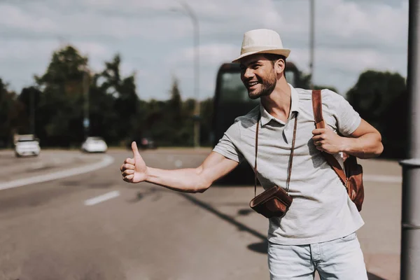 Young Tourist with Backpack Hitchhiking on Road. Casually dressed Handsome Man with Camera on Neck trying to Catch Car on Highway. Tourism and People Concept. Active Lifestyle. Summer Vacation