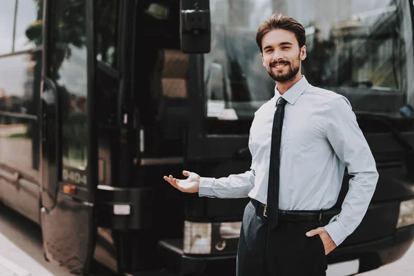 Young Smiling Businessman Standing in front of Bus. Confident Attractive Man wearing White Shirt and Black Tie Standing in front of Tour Bus Inviting to come in. Successful Professional Employee