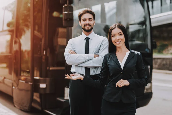 Young Smiling Business People Standing next to Bus. Confident Attractive Man with Crossed Arms and Beautiful Woman Standing in front of Tour Bus Inviting to come in. Successful Professional Employees