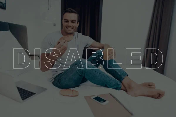 Disabled Man. Man is Sitting on Bed. Laptop Computer Lying on Bed. Smartphone and Notebook Lying on Bed. Man is Happy and Smiling. Man Eating a Cake. Man Drinking Juice in Cup. Man Located in Bedroom.