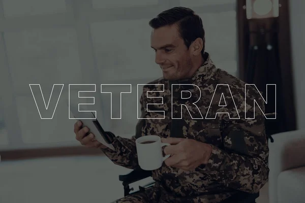 Military Veteran. Disabled Man in a Wheelchair. Man Watching Video on Tablet PC. Man is Soldier. Soldier in Military Uniform. Man Drinking Coffee in Cup. Man Smiling. Man Located in the Living Room.