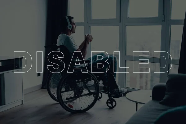 Disabled Man in a Wheelchair. Man Sits in Front of Window. Man Looks into Window. Man Listening Music on Headphones. Man is Thinking. Man Drinking a Coffee. Man Located in the Living Room.