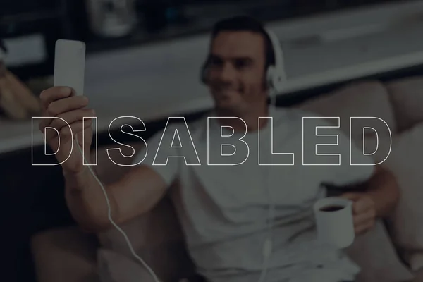 Disabled Man. Man is Sitting on Sofa. Man Wearing a Headphones. Man Listening a Music. Music on Mobile Phone. Man is Smiling. Man Drinking Coffee in Cup. Man Located in the Living Room. Blur Image.