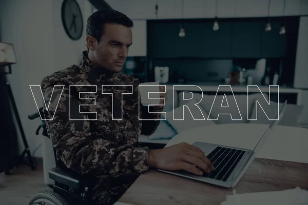 Military Veteran. Disabled Man in a Wheelchair. Man Sitting at Table. Man Using s Laptop. Man is Soldier. Soldier in Military Uniform. Man Drinking Coffee in Cup. Man Located in the Living Room.