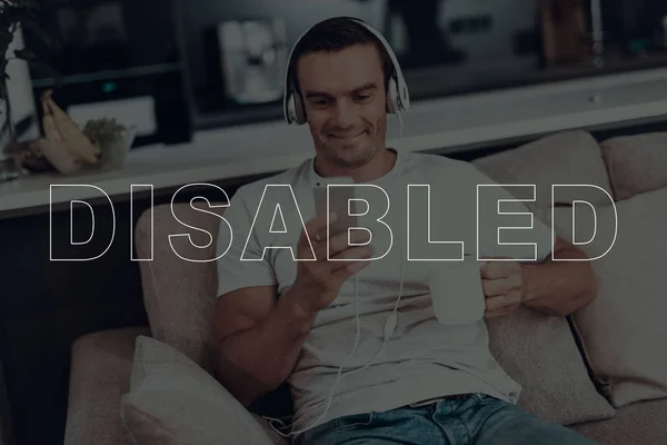 Disabled Man. Man is Sitting on Sofa. Man Wearing a Headphones. Man Listening a Music. Music on Mobile Phone. Man Happy and Smiling. Adult Brunette Man. Man Located in the Living Room.