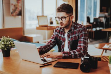 Portre yakışıklı Freelancer dizüstü bilgisayarda çalışıyor. Yakışıklı beyaz Hipster fotoğrafçı gözlük oturur masaya gadget'lar ve kapalı klavye ile giyer. Mesafe iş kavramı