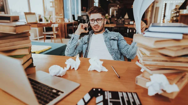 Text Writer Talking on Phone and Working Indoors. Portrait of Handsome Journalist or Screenwriter Artist Sitting at Desk With Two Big Pile of Books and Movie Clapper Board Using Laptop.