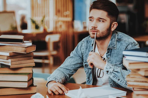 Concept of Young Text Writer Working Indoors. Handsome Hardworking Freelance Screenwriter Scenarist Sitting at Table Near Two Big Stacks of Books Writing Article, Novel, Play on Paper Sheets.