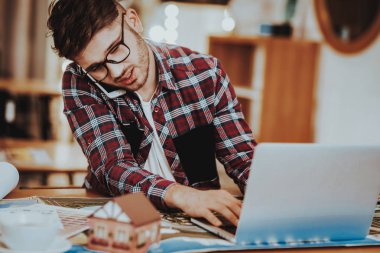 İş adamı inşaat üstünde Laptop görüşmeler telefon ile meşgul. Portre portre yakışıklı ciddi beyaz Freelancer Smartphone konuşmayı işyerinde oturuyor. Uzak iş kavramı