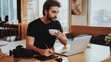 Mutlu adam, dizüstü bilgisayarda çalışırken kahve içiyor. Portre yakışıklı sakallı Grafik Tasarımcısı kullanır dijital tablet kalemi ile İşyeri rötuş fotoğraf ahşap masada oturur. Freelancing kavramı