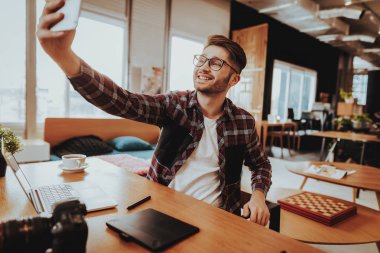 Serbest meslek sahibi olun İşyerinde çalışırken Selfie. Gözlük kullanır Smartphone ön kameraya bakarak şık sakallı gülümseyen yakışıklı beyaz adam portresi kapalı masada oturur.