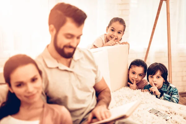 Happy Cute Parents Reading Book while Kids Playing. Naughty Children Annoying Parents. Three Kids Having Fun while Dad and Mom Having Rest, Lying on Sofa and Reading Books. Family Concept.