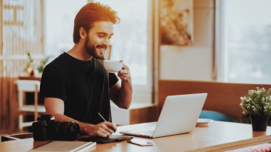 Gülümseyen adam dizüstü bilgisayarda çalışırken çay içer. Portre yakışıklı sakallı grafik tasarımcı Stylus oturur, işyerinde ahşap masa ile dijital Tablet rötuş fotoğraf kullanır. Freelancing kavramı