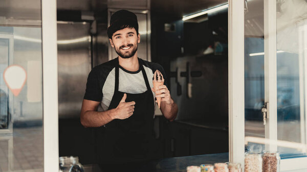 Young Bearded Man in Apron Standing in Food Truck. Ice Cream in Hand. Street Food Concept. Food in Town. Selling Snacks. Guy in Cap. Black Arpon. Working Outdoor. Man in T-shirt. Standing Guy.